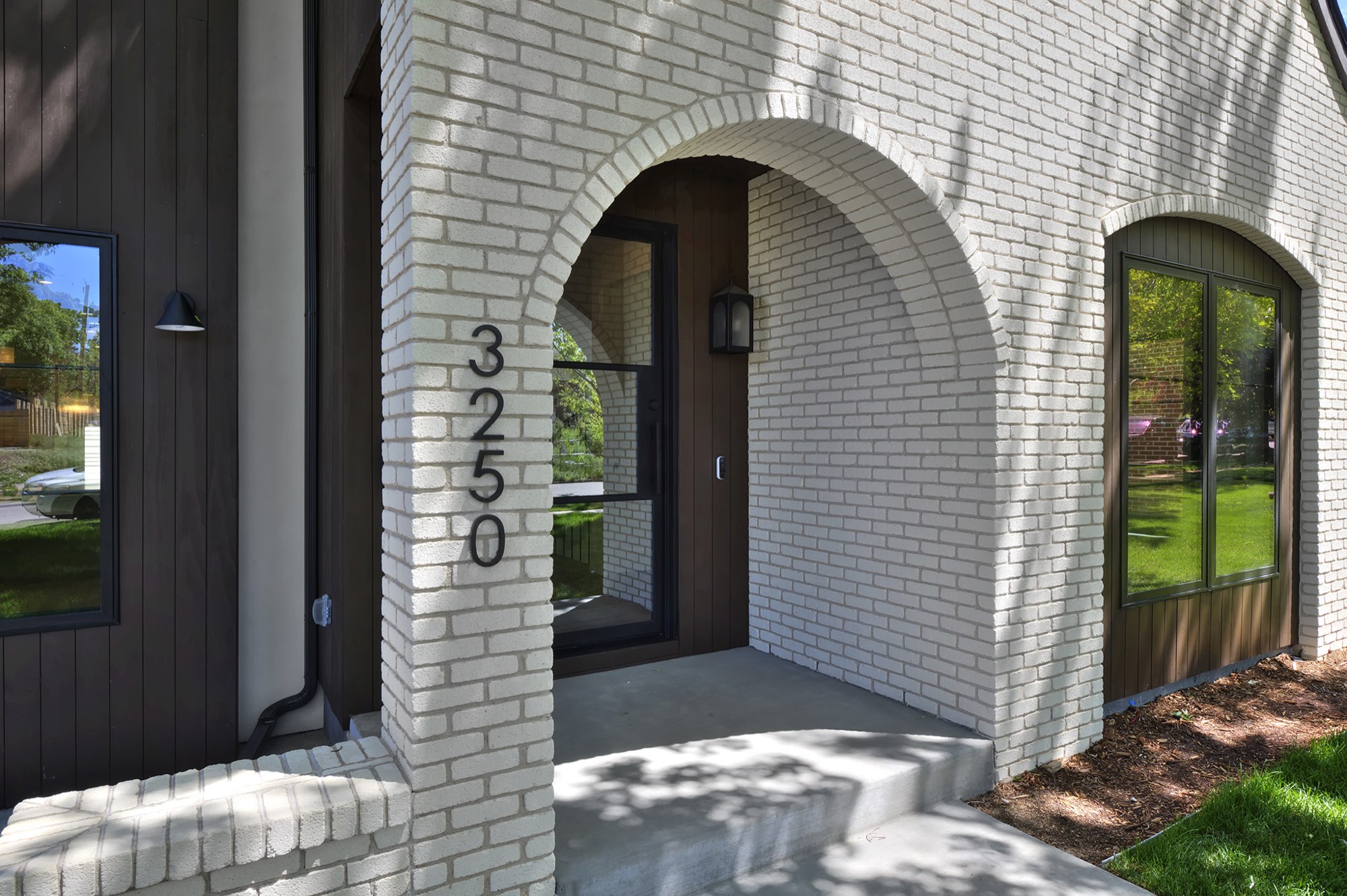Arched brick entry with the house number 3250 set into the white brick beside dark vertical siding and a black-framed arched window