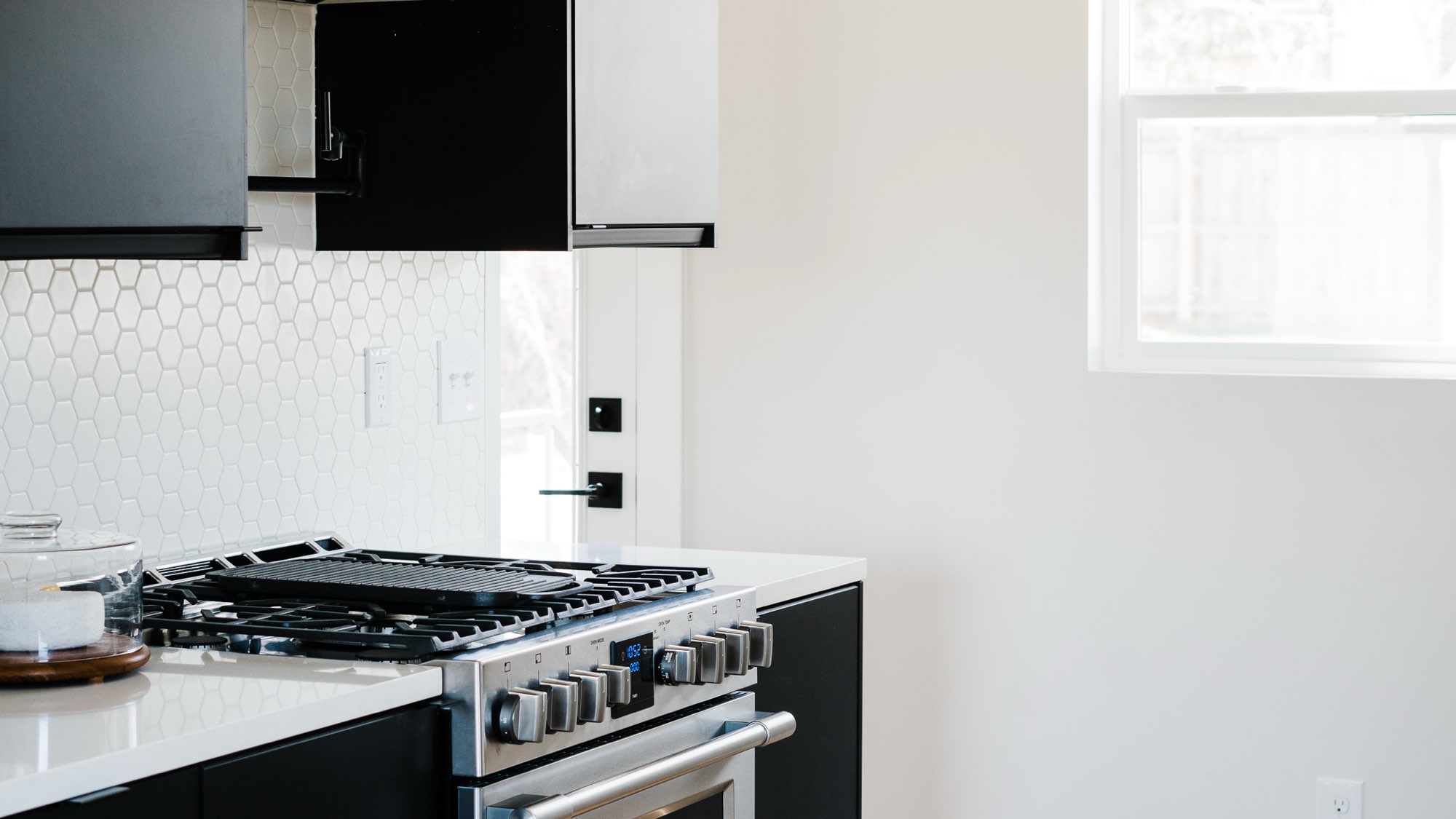 Kitchen with a stainless gas range, white hexagon tile backsplash, white counters and black cabinet doors