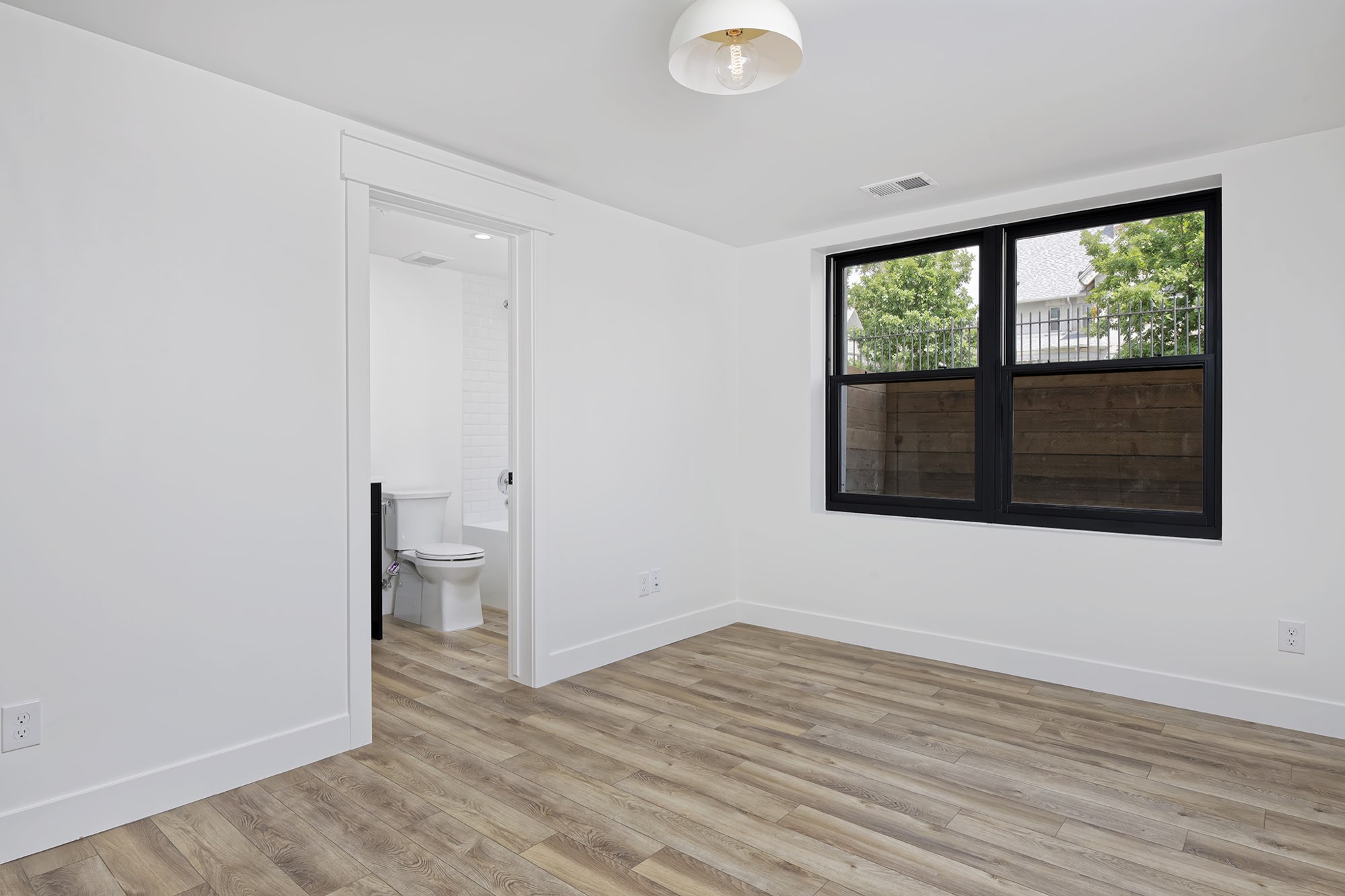 Lower-level bedroom with light wood-look flooring, a black-framed window and a bathroom doorway