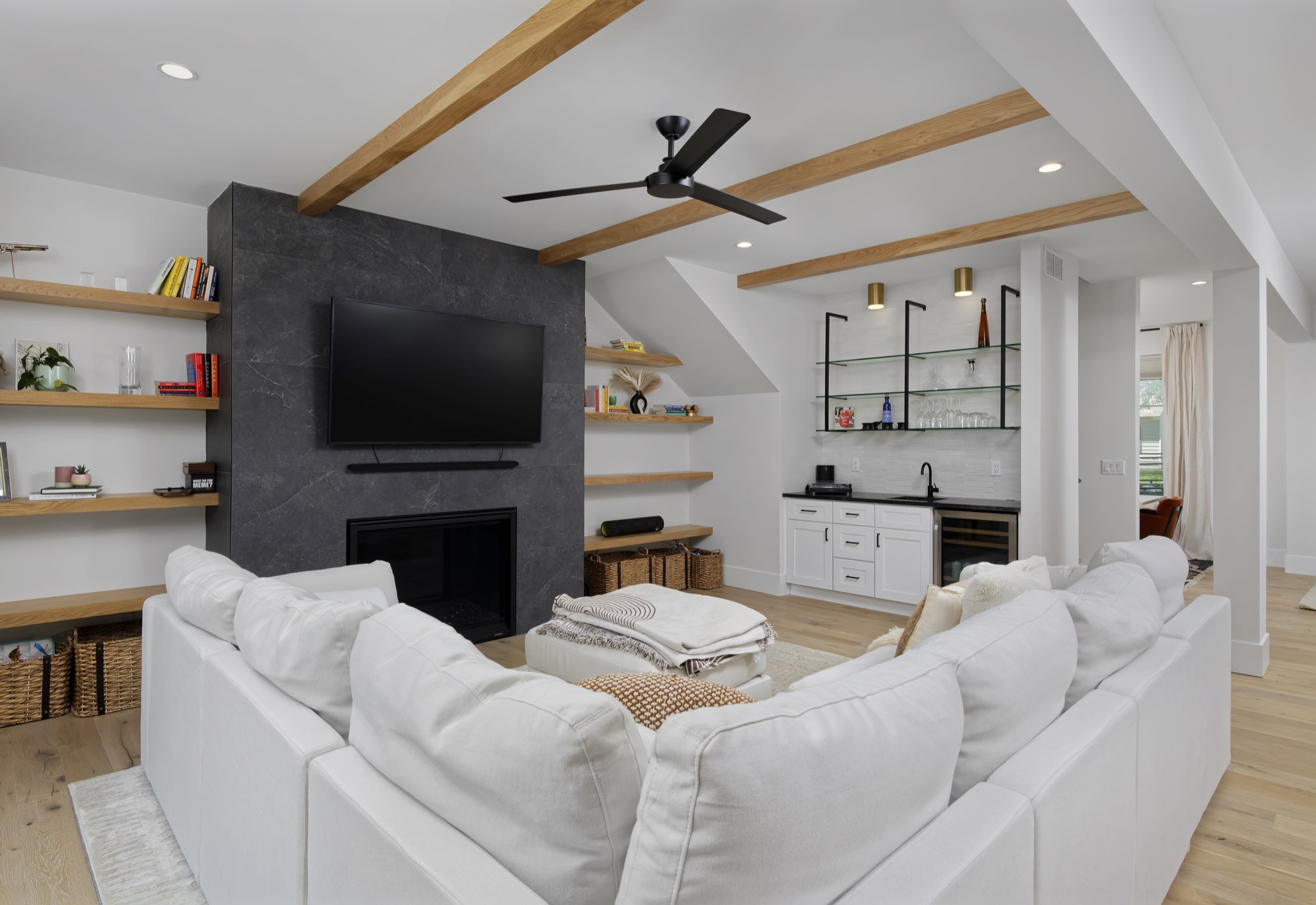 Living room with a dark stone fireplace, wood ceiling beams, floating shelves and a wet bar in the corner