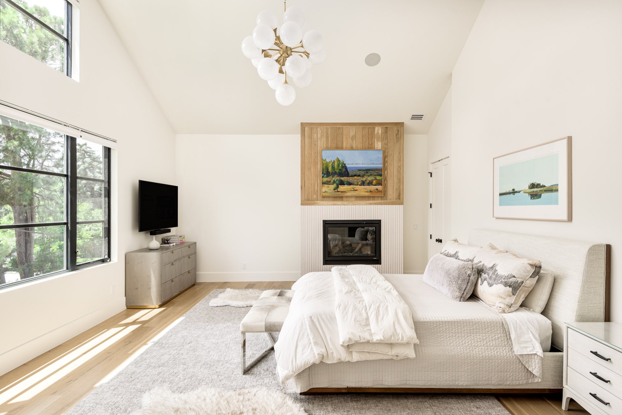 Bedroom with a vaulted ceiling, a fireplace in a wood-clad wall, a cluster chandelier and tall black-framed windows