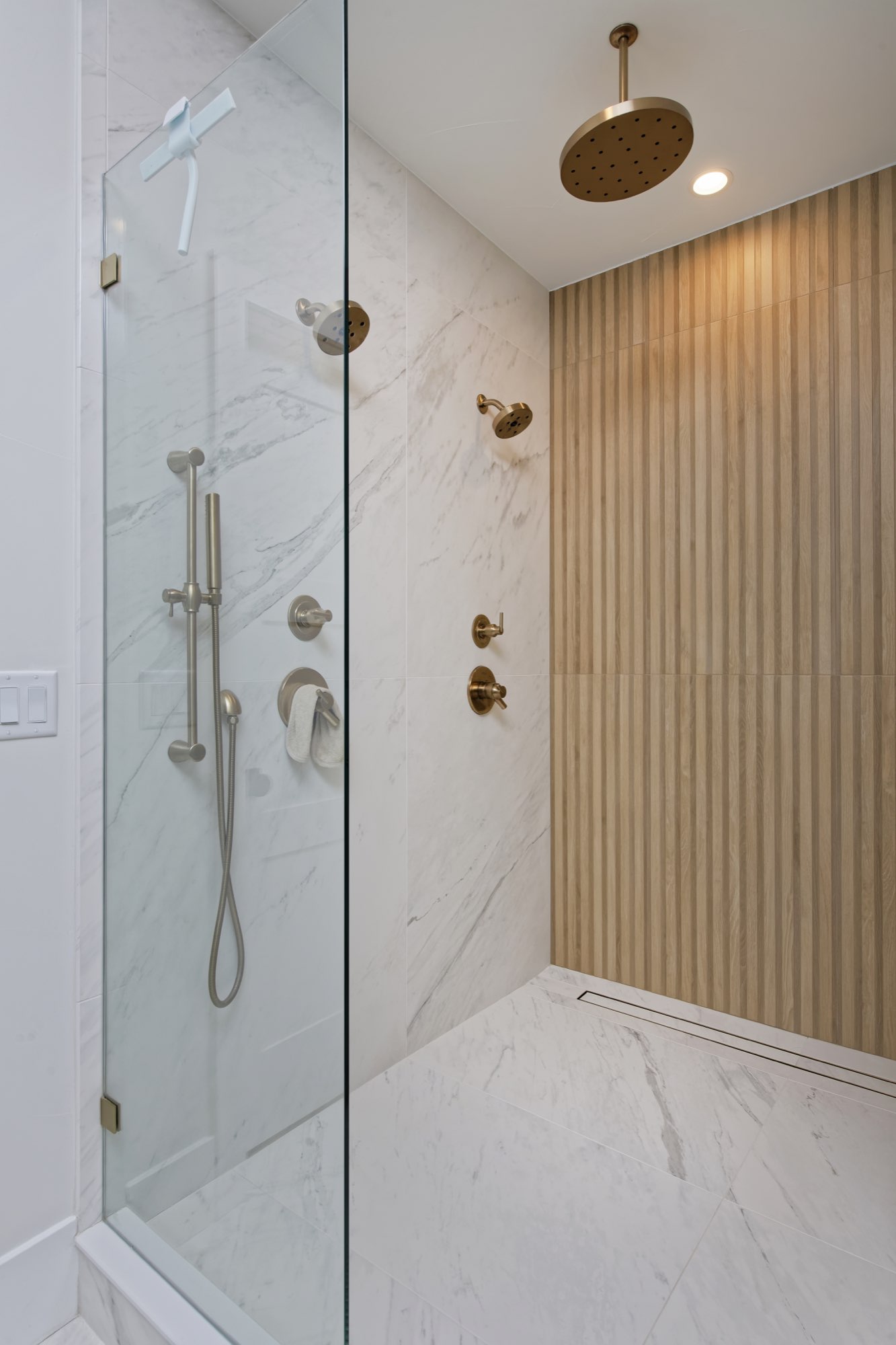 Walk-in glass shower with white veined tile, a wood slat wall, a rain head and brass fixtures